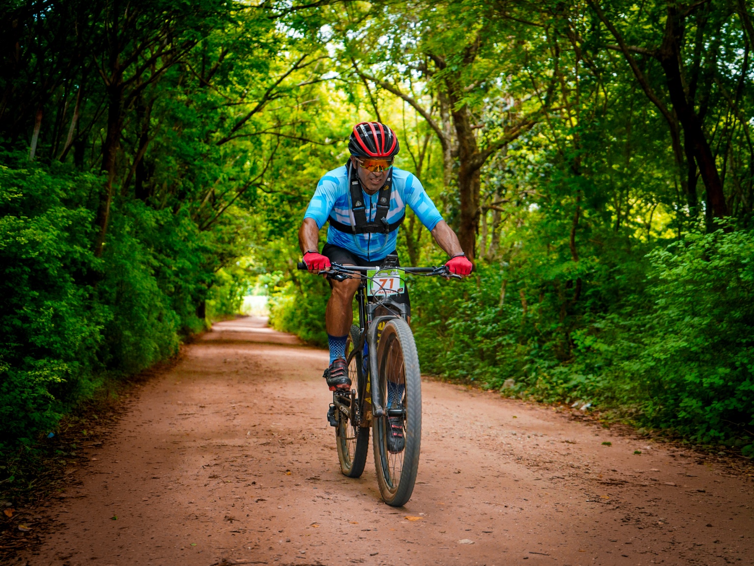 Ciclista de camisa azul pedalando em uma estrada de terra cercada por mata verde.