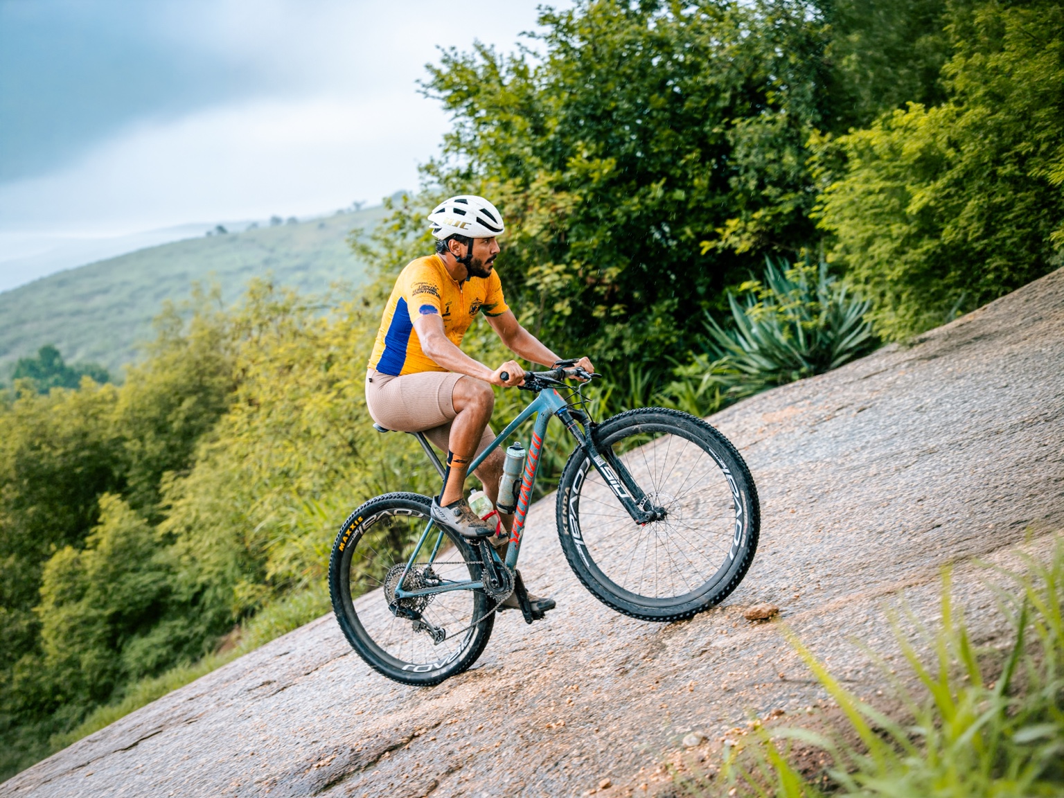 Ciclista de camisa amarela subindo uma trilha de pedra, com serras e vegetação ao fundo.