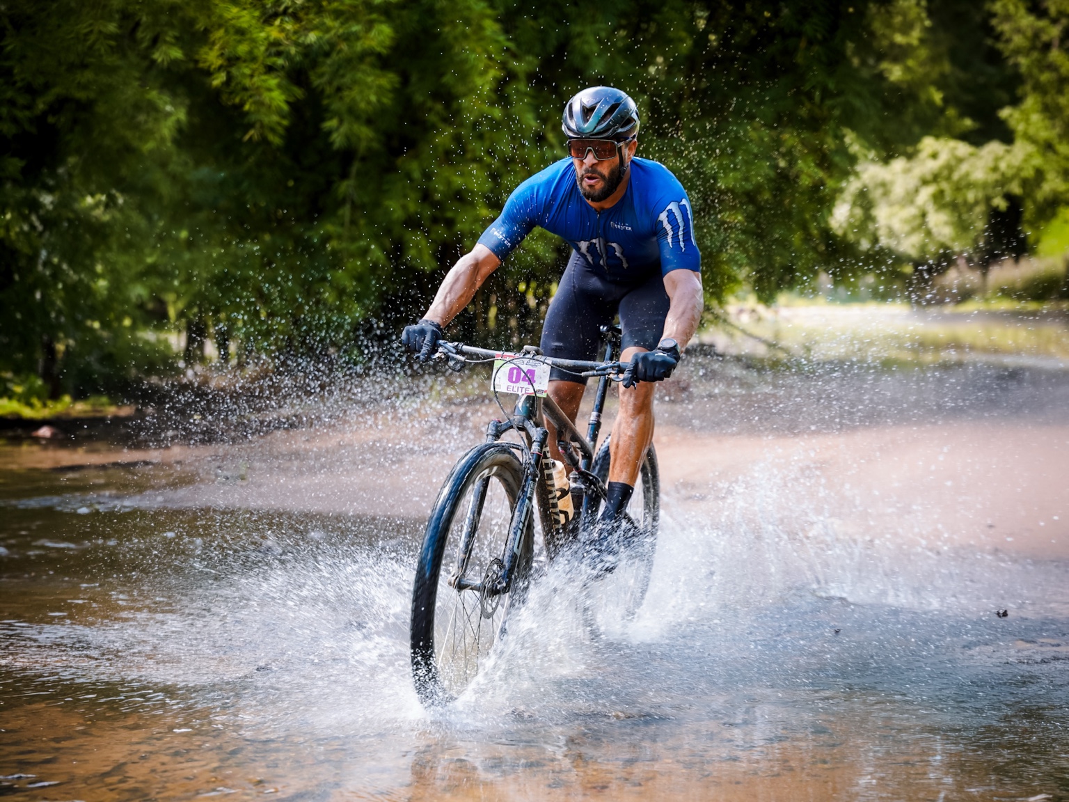 Ciclista de camisa azul atravessando a água em uma trilha cercada por vegetação.
